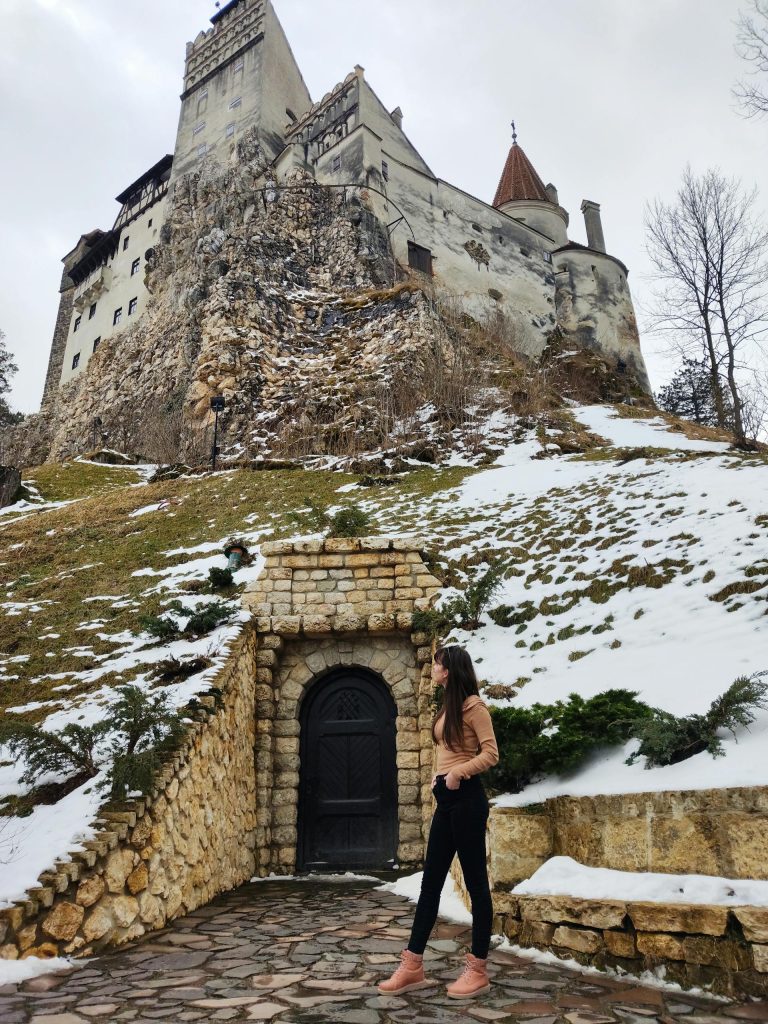 Snow-covered Bran Castle with a woman standing near the entrance, showcasing Romanian winter beauty.