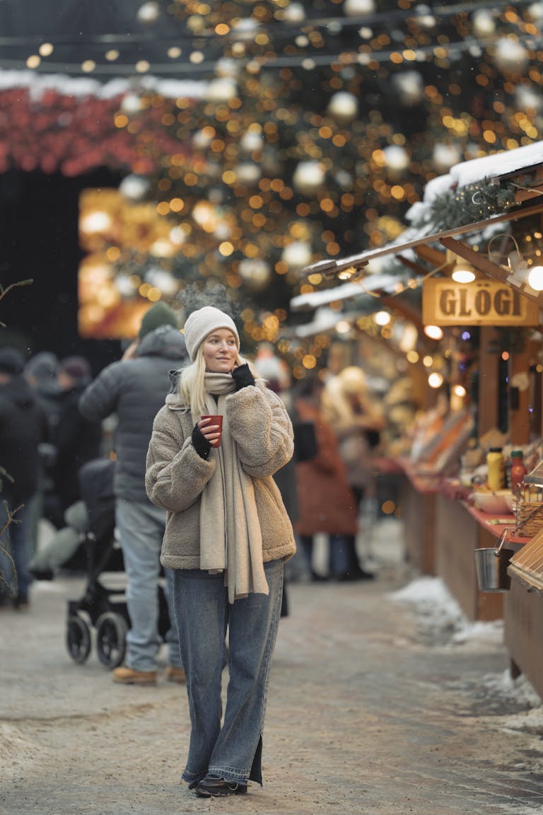 A woman stands warmly dressed holding a drink at a vibrant Christmas market.