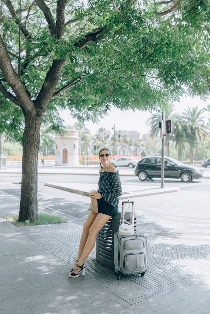 Fashionable woman sitting with luggage on sunny city street, enjoying travel.